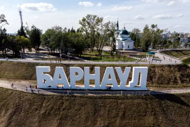 Photo of Close up to the hill with the letters spelling out Russian city's name Barnaul, a park and a church at the background. City's landmark and architrctural brand. Popular recreational place among citizens