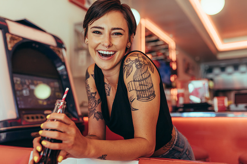 Smiling woman with tattooed arms standing in a diner. Woman standing beside a gaming machine holding a soft drink.