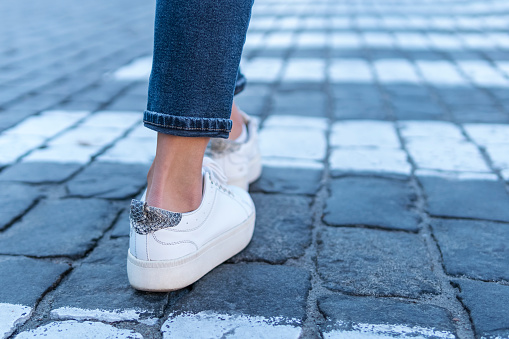 Female legs in white sneakers at a pedestrian crossing, close-up, rear view