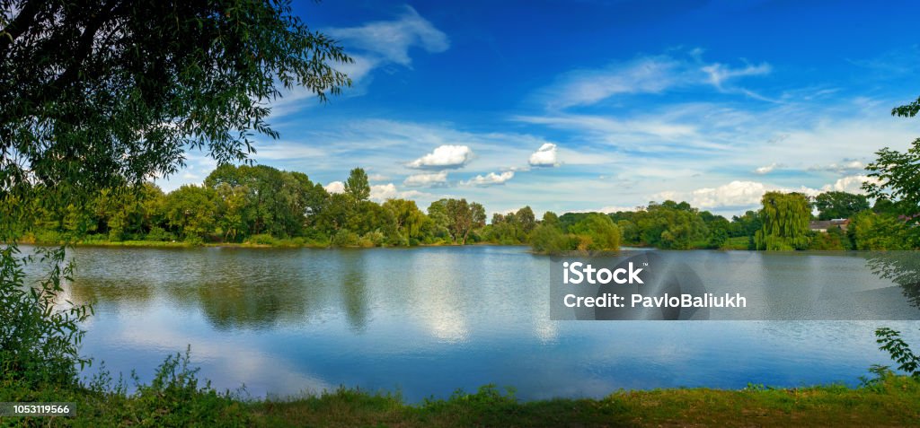 Tranquil landscape at a lake, with white clouds sky and the trees reflected in the clean blue water Tranquil landscape at a lake, with the vibrant sky, white clouds and the trees reflected in the clean blue water Riverbank Stock Photo