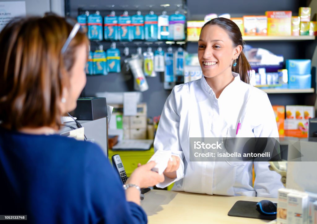 Smiling attractive young redhead pharmacist handing over prescribed medicines to an elderly female patient, view over the clients shoulder of the pharmacist Female pharmacist staff counseling customer about drugs usage in modern pharmacy Pharmacy Stock Photo