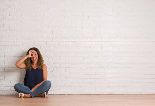 istock Middle age hispanic woman sitting on the floor over white brick wall doing ok gesture with hand smiling, eye looking through fingers with happy face. 1053159782