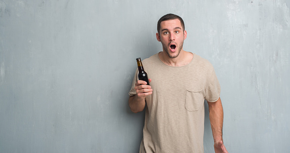 Young caucasian man over grey grunge wall holding bottle beer scared in shock with a surprise face, afraid and excited with fear expression