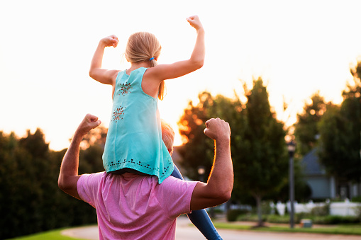 Little girl on top of fathers shoulders holding arms up as in showing muscle.