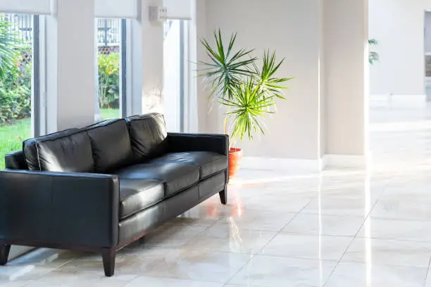 Black leather couch, sofa with green potted palm tree plant in pot with tiles, tiled floor in hall, room, lobby of residential, condo, condominium house, building, complex with bright, natural light