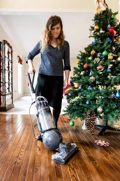 Young woman cleaning with vacuum cleaner hardwood floor after New Year needles from Christmas tree in living room