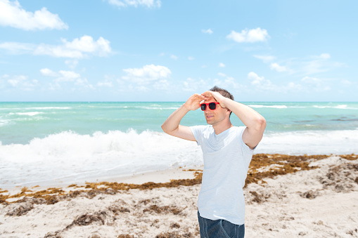 Young man in sunglasses standing on sand, sandy beach in Miami, Florida by ocean, sea water, waves on sunny day with blue sky, seaweed, two hands holding on forehead