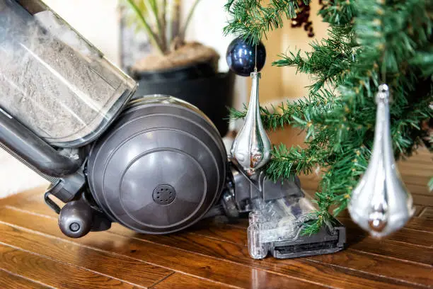Closeup of upright vacuum cleaner head cleaning, vacuuming under Christmas Tree needles with New Years ornaments on hardwood wooden floor