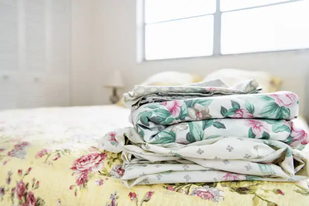 Stack of bedroom linen with floral, flower pattern on top of bed with quilt, quilted comforter, duvet, bright natural light from window, lamp, closet in background