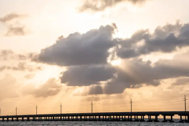 Overseas highway bridge road at Bahia Honda key state park during dramatic sunset with sun behind clouds, god rays, power electricity lines, water, waves