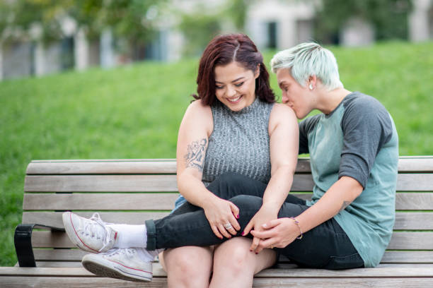 Canoodling on a park bench A lesbian or LGBTQ couple sit on a park bench. One has her legs on her partner's lap, and is kissing her shoulder and holding her hand. Her partner is smiling and blushing. canoodling stock pictures, royalty-free photos & images