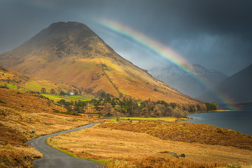 Bright rainbow soaring over the blue shore of Wast Water and Yewbarrow overlooked by the snowy peaks of Great Gable and Scafell in this picturesque panoramic vista of the Lake District National Park, Cumbria, UK.