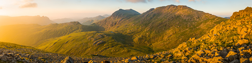 Golden light of sunrise filling the green mountain valleys high in the Lake District National Park overlooked by the iconic rocky summits of the Langdale Pikes and Bow Fell towards Coniston and Windermere, Cumbria, UK.