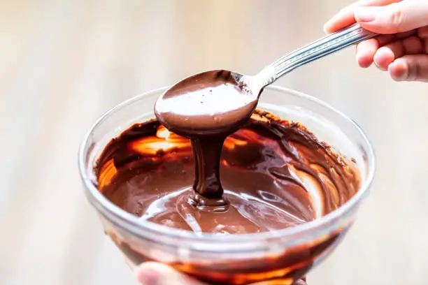 Closeup of woman hand holding spoon, mixing, flowing, stirring raw vegan liquid, creamy, smooth chocolate mousse, cream, icing in bowl, plate, dish