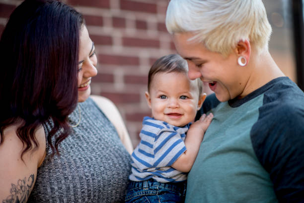 Young same sex couple adoringly smile at their baby boy Same sex couple hold their baby son close to their chest as they smile at him. They are hugging their son tight as he smiles looking out into the distance. homosexual couple stock pictures, royalty-free photos & images