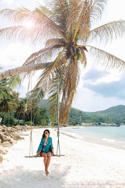 Young woman enjoying the tropical paradise on Koh Phangan island in Thailand Pastel vintage toned image of a young brunette woman, enjoying her travel and vacation on the beautiful island of Koh Phangan, in the Gulf of Thailand. She is sitting on a swing by the palm tree, having a lazy day off. thailand beach stock pictures, royalty-free photos & images