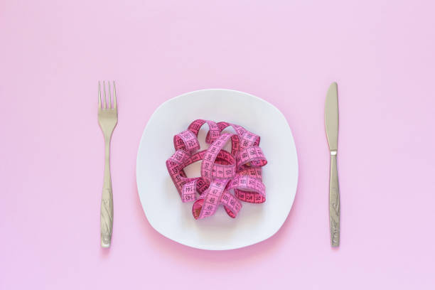 Pink measuring tape lying on plate in the form of spaghetti, knife and fork on pink background stock photo