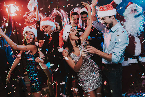 Happy Young People Dancing on New Year Party. Santa Claus. People in Red Caps. Happy New Year Concept. Glass of Champagne. Celebrating of New Year. Young Woman in Dress. Men in Suits.