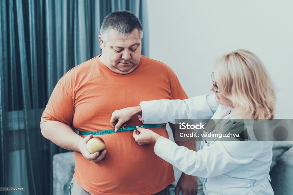 Fat Man with Doctor in White Coat in Gray Room. Fat Man with Doctor in White Coat in Gray Room. Woman with Tailors Centimeter. Diet and Healthcare Concept. Man with Bulimia. Unhealthy Lifestyle Concept. Man with Overweight. Patient with Stomach. Overweight Stock Photo