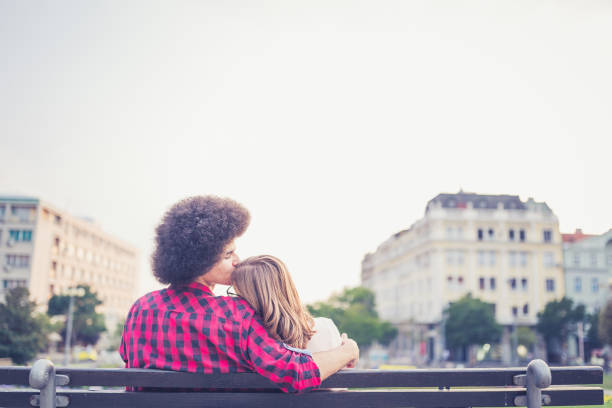 Ellis Island, New York, NY, USA – September 21th, 2023: A middle-aged man and woman wearing sneakers hold each other while sleeping on a bench in the buildings on Ellis Island New York. The man has a backpack on his lap.