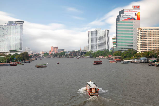 View over Bridge on BTS Station with Chao Phraya river in. Several skyscrapers and to a moving boat in the center. Traffic on the river. Blue sky with fluffy white clouds but some haze.