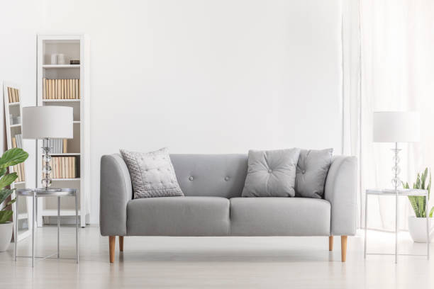 istock Lamp on silver table next to grey sofa with pillows in white living room interior with plant. Real photo 1053407140
