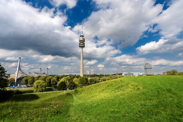MUNICH, GERMANY - SEPT 8, 2018:  Olympiapark (Olympic Park), Olympiasee (Olympic Lake) and Olympiaturm (Olympic Tower) in Munich, was constructed for the 1972 Summer Olympics. On background the headquarters of the BMW in Munich, Bavaria, Germany, Europe. The Park continues to serve as a venue for cultural, social, and religious events, such as events of worship, is administered by Olympiapark München GmbH, a holding company fully owned by the state capital of Munich. Tourists and locals stroll in the park.