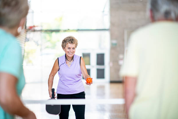 Playing Pickleball A group of seniors are playing pickleball in a fitness center. One woman is about to serve. pickleball stock pictures, royalty-free photos & images