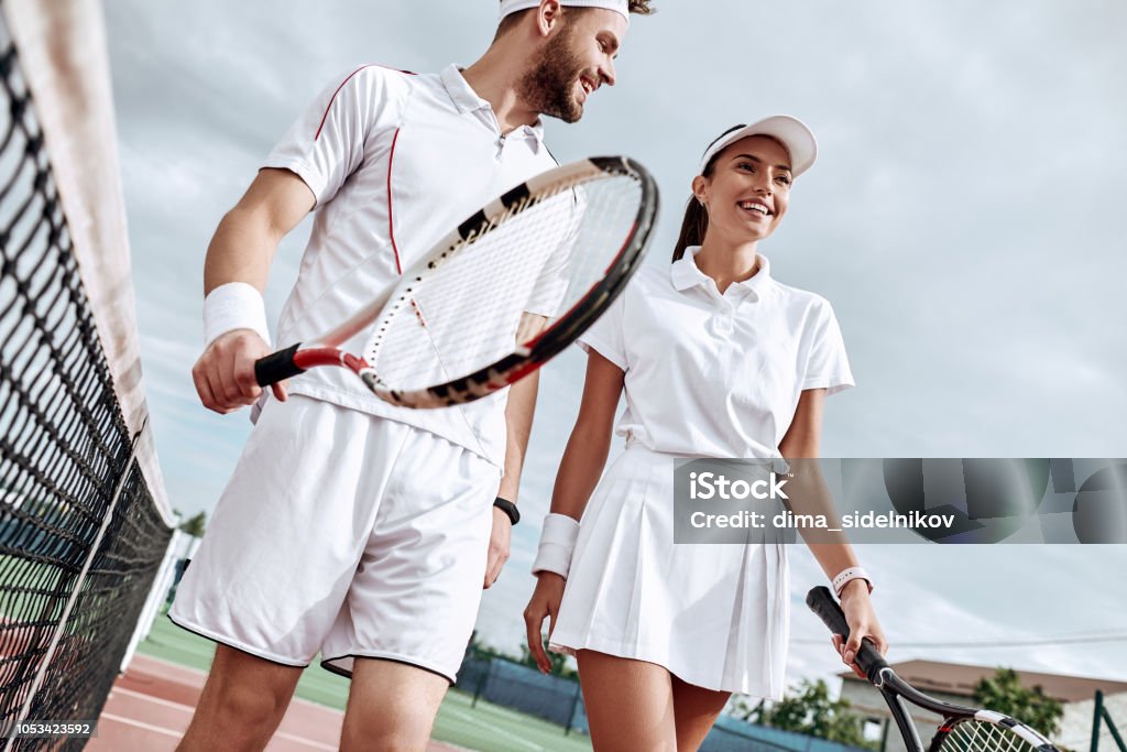 Aproveitando-se de passar tempo na quadra. Lindo casal jovem caminhando na quadra de tênis com sorriso. - Foto de stock de Tênis - Esporte de Raquete royalty-free