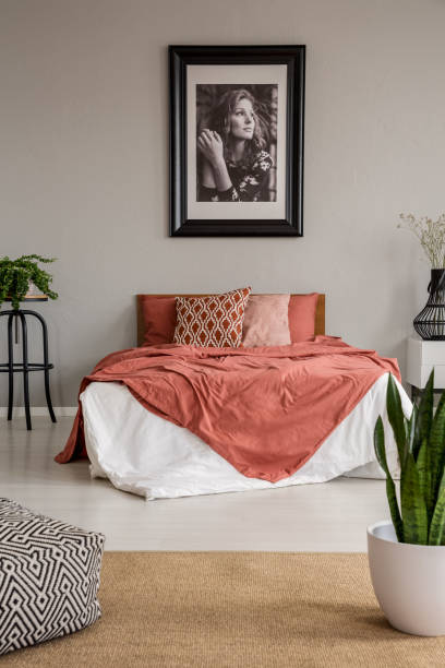 istock Poster on grey wall above bed with red sheets in bedroom interior with pouf and plants. Real photo 1053430352