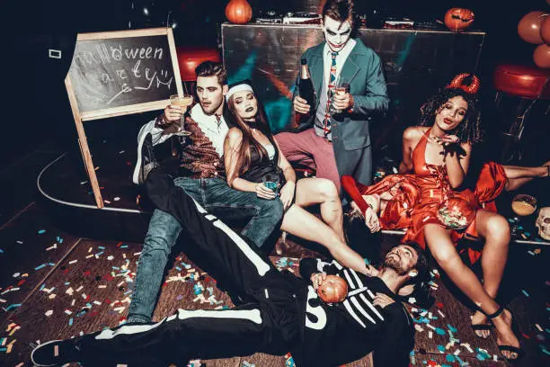 Drunk Young People in Costumes Resting after Party. Group of Young Friends Wearing Costumes Resting after Halloween Party by lying on Floor of Nightclub and Drinking. Nightlife Concept
