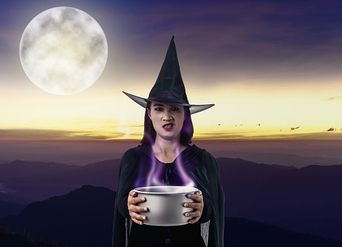 Portrait of woman in black Scary witch halloween costume holding spooky witch's cauldron smoke coming out with moonlight. landscape mountain and sea of mist is background