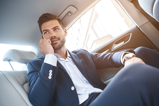 Smiling business man talking on smart phone and sitting in backseat of the car