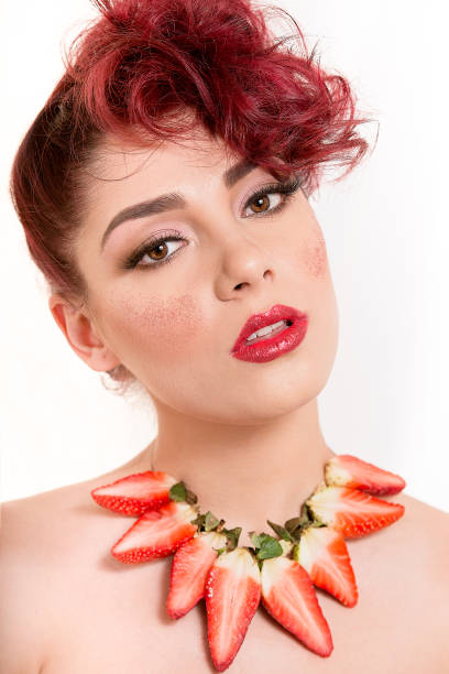 istock Beautiful Caucasian redhead woman looking sensual with open mouth and tilted head, having necklace of strawberries, red glitter on the cheeks and bare sholders, on white background 1053486310