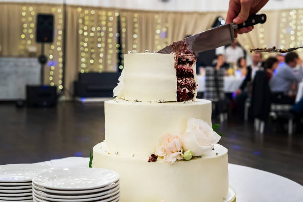 istock Cutting the first tier of white mastic wedding cake in banquet hall background. Front view at table with a beautiful wedding cake and side plates for serving guests. Dividing wedding cake into parts. 1053497598