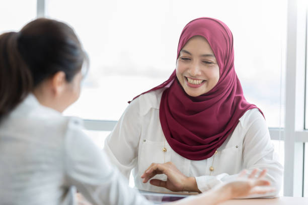 Malaysian businesswoman discusses project with coworker A smiling young Malaysian businesswoman sits across her office desk from an unrecognizable coworker.  She looks down as they discuss a project. malaysia office workers stock pictures, royalty-free photos & images