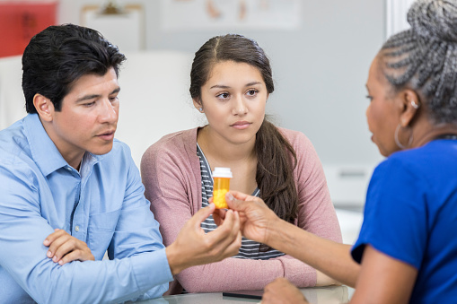 Serious senior African American female healthcare provider discusses drug side effects with a teenage female patient and the girl's father.