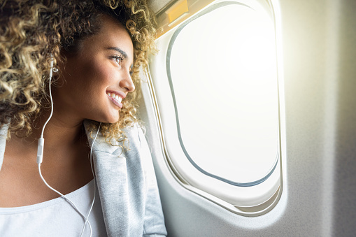 An excited young businesswoman smiles as she looks out the window.  She is sitting in the window seat of a commercial airliner.  She also wears a pair of earbuds.