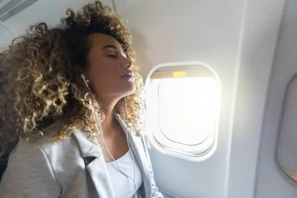 A young woman leans her head back and closes her eyes as she sits in the window seat of a commercial airliner.  She also listens to music on her earbuds.
