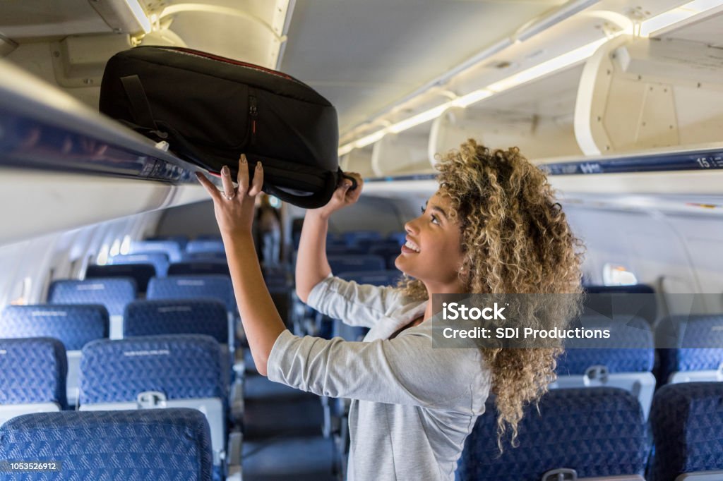 Young woman places luggage in airline overhead bin A smiling young businesswoman stands in the aisle of a commercial airliner and places her carry on bag in the overhead bin. Carry-On Luggage Stock Photo