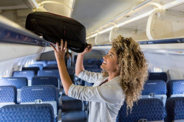 Young woman places luggage in airline overhead bin A smiling young businesswoman stands in the aisle of a commercial airliner and places her carry on bag in the overhead bin. hand luggage stock pictures, royalty-free photos & images
