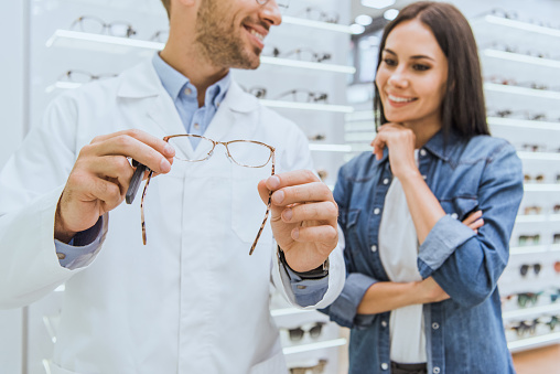 partial view of happy male oculist helping female to choose pair of eyeglasses in ophthalmic shop