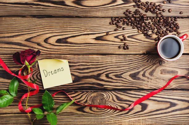 sueños de inscripción en papel al lado de rosa roja con cinta y taza de café junto a granos de café en la mesa de madera vieja. vista superior con espacio de copia. concepto de día de san valentín - coffee bean coffee flower ribbon fotografías e imágenes de stock