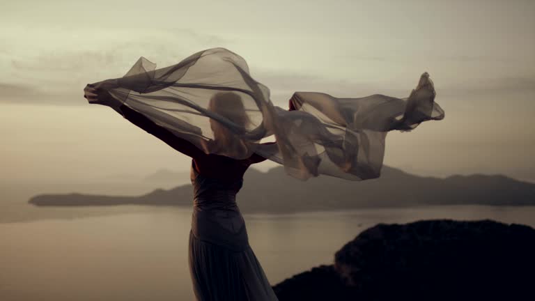 Young woman in long summer dress flying in the wind standing on a rocky shore