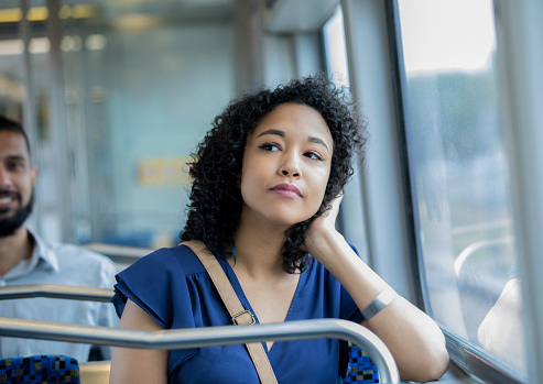 Young adult businesswoman is resting her head in her hand and daydreaming while sitting in commuter train car. Professional is using public transportation to reach her destination.