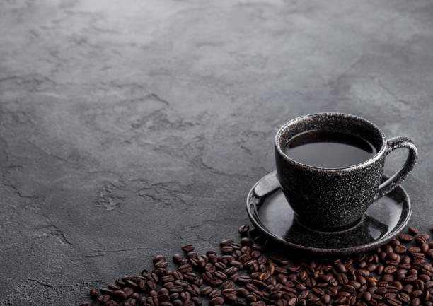 Black coffee cup with saucer and fresh coffee beans on black stone kitchen table background.