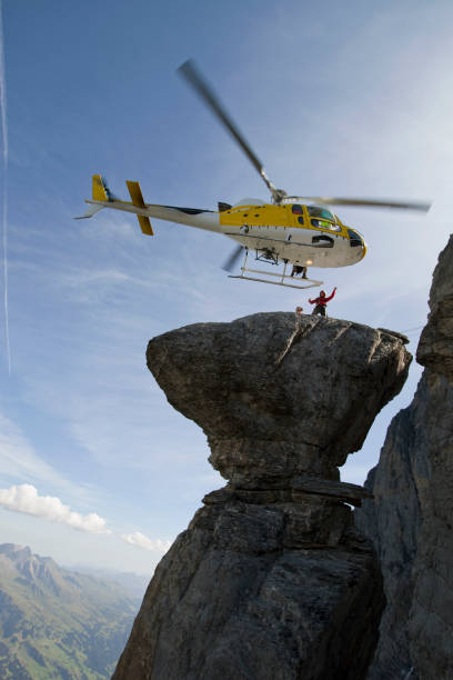 Yellow helicopter on background of high Alps mountains wth green forest under blue cloudy sky.