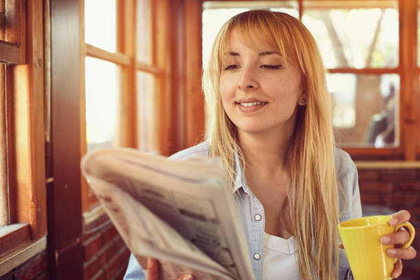 beautiful young woman reading newspaper at a cafe and holding coffe cup in hand with positive expression - job search newspaper coffee shop cafe imagens e fotografias de stock
