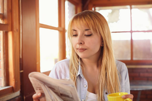 beautiful young woman reading newspaper at a cafe, holding coffe cup in hand and searching job - job search newspaper coffee shop cafe imagens e fotografias de stock