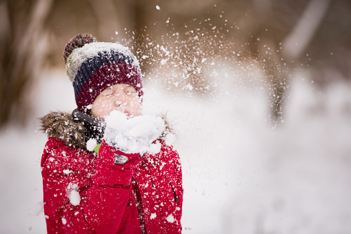 Portrait of cute kid boy blowing snow from his hands on a winter day. Child playing outdoors. Lifestyle concept